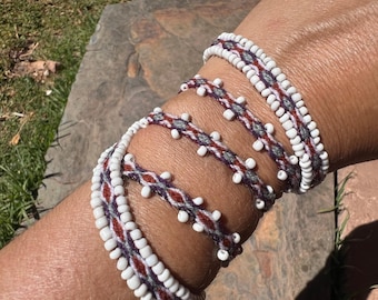 May include: A multi-wrap bracelet featuring a woven cord in shades of dark red, grey, and white, accented with small white seed beads. The design incorporates a repeating diamond pattern along the center, bordered by rows of white beads.