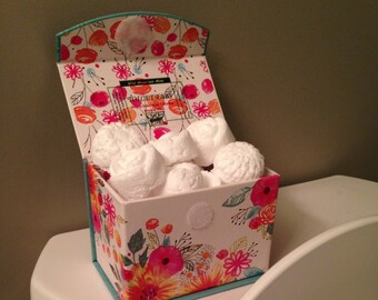 May include: A floral patterned box filled with white toilet bowl freshener drops. The box is sitting on a white toilet.