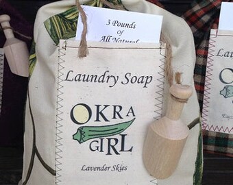 May include: A white fabric bag with a brown twine tie, filled with laundry soap. The bag has a label that says "Laundry Soap Okra Girl Lavender Skies" and "3 Pounds of All Natural". A small wooden scoop is attached to the bag.