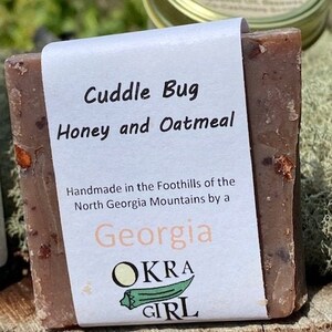 May include: A brown bar of soap with the text "Cuddle Bug Honey and Oatmeal" on a white label. The label also includes "Handmade in the Foothills of the North Georgia Mountains by a Georgia OKRA GIRL" in black and light orange text.