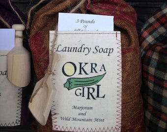 May include: A burlap bag of laundry soap with a red and gold plaid pattern, tied with twine. The label reads "Laundry Soap OKRA GIRL" with a green okra pod graphic. The bag contains 3 pounds of soap.