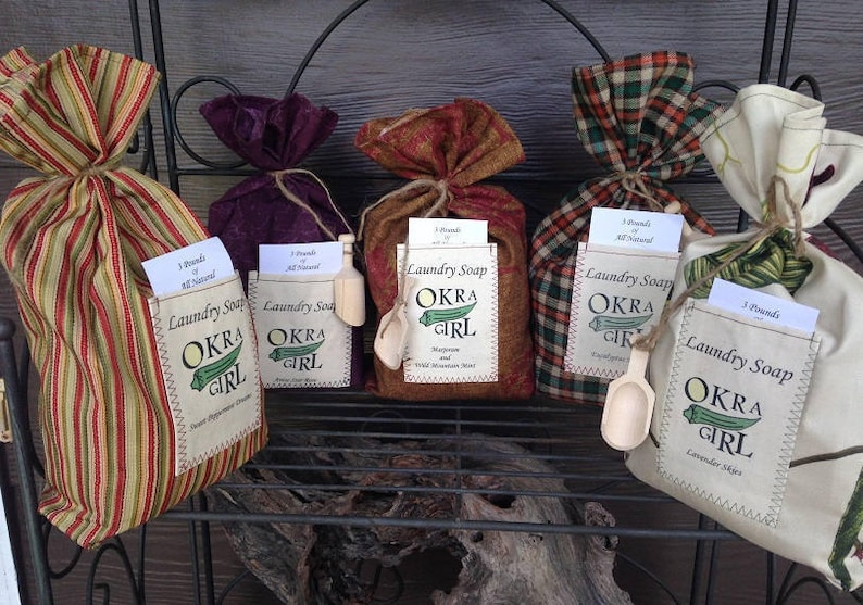May include: Four bags of laundry soap with a label that says "Okra Girl" in a variety of fabric patterns. The bags are filled with laundry soap and have a small wooden scoop attached to each bag.