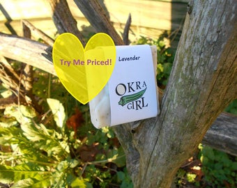 May include: A white bar of soap with a label that says "Okra Girl" and "Lavender". The soap is sitting on a branch with a yellow heart that says "Try Me Priced!" in pink text.