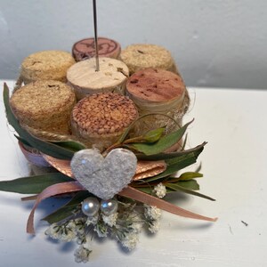 May include: A small, rustic, cork-based picture frame with a white heart and pearl accents. The frame is decorated with burlap, greenery, and a peach-colored ribbon.
