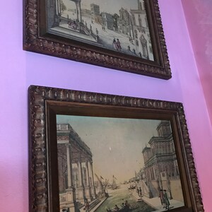 May include: Two framed antique prints of cityscapes. The top print shows a view of a city square with buildings and people. The bottom print shows a canal with boats and buildings on either side.