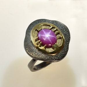 Puede incluir: Un anillo de plata con un zafiro estrella rosa engastado en un bisel de oro. El anillo tiene una banda texturizada y un diseño único.