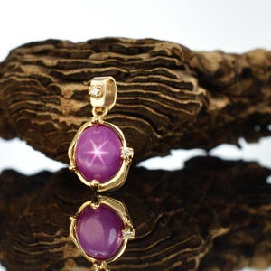 May include: A gold pendant featuring a pink star sapphire gemstone with a star-like pattern. The pendant is set in a gold frame with small diamonds on the sides.