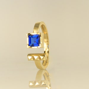Op de afbeelding: Een gouden ring met een blauwe vierkante edelsteen in het midden. De ringband is getextureerd met kleine driehoeken.