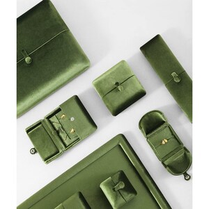 May include: A collection of olive green velvet jewelry storage cases and boxes in various shapes and sizes. The set includes rectangular, square, and elongated containers, some featuring small button closures and internal compartments for jewelry.