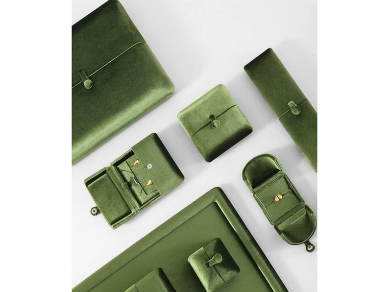 May include: A collection of olive green velvet jewelry storage cases and boxes in various shapes and sizes. The set includes rectangular, square, and elongated containers, some featuring small button closures and internal compartments for jewelry.