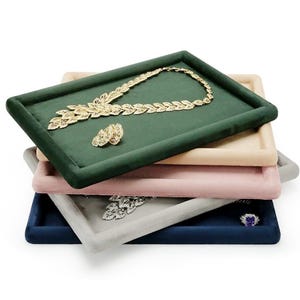 Può includere: Una pila di vassoi espositivi per gioielli in vari colori: verde, beige, rosa, grigio e blu navy. Una collana e un anello d'oro poggiano sul vassoio verde. Altri vassoi contengono gioielli argentati e viola.