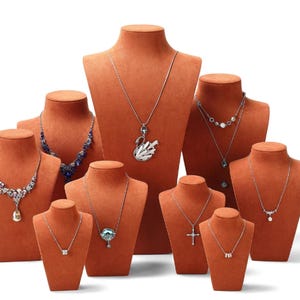 Puede incluir: Una colección de collares exhibidos en bustos de terciopelo naranja. Los collares presentan varios diseños, incluyendo colgantes, piedras preciosas y cadenas delicadas. Las joyas se presentan sobre un fondo blanco, resaltando los detalles.