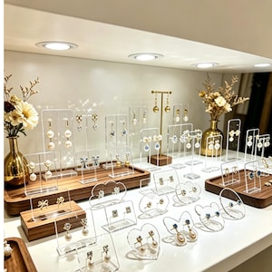 May include: A collection of various earrings displayed on clear acrylic stands and wooden trays. The earrings feature pearls, blue stones, and metallic bow designs. Two gold vases with dried flowers are placed on the sides of the display.