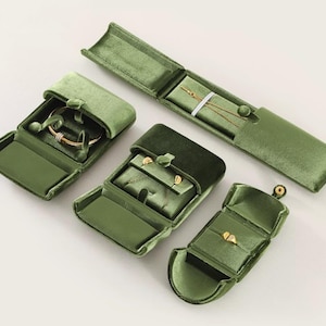 May include: A collection of four olive green velvet jewelry boxes in various shapes and sizes. One long rectangular box holds a thin gold chain, while the other three square and rectangular boxes contain gold rings and small gold stud earrings.