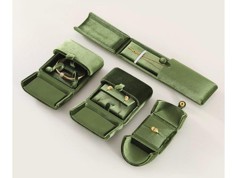May include: A collection of four olive green velvet jewelry boxes in various shapes and sizes. One long rectangular box holds a thin gold chain, while the other three square and rectangular boxes contain gold rings and small gold stud earrings.
