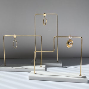 Könnte beinhalten: Drei goldene Ohrringständer aus Metall mit weißen Marmorsockeln. Jeder Ständer hat einen rechteckigen Sockel und eine vertikale Stange mit einer horizontalen Stange oben. Die Ständer sind in einer Reihe angeordnet, wobei der mittlere Ständer etwas höher ist als die beiden anderen.