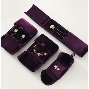 Op de afbeelding: Een bovenaanzicht toont verschillende open, dieppaarse fluwelen sieradendoosjes. Elk doosje bevat goudkleurige sieraden, waaronder oorbellen, een armband en kettingen. De doosjes zijn gerangschikt op een witte ondergrond.