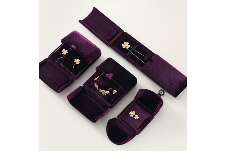 Op de afbeelding: Een bovenaanzicht toont verschillende open, dieppaarse fluwelen sieradendoosjes. Elk doosje bevat goudkleurige sieraden, waaronder oorbellen, een armband en kettingen. De doosjes zijn gerangschikt op een witte ondergrond.