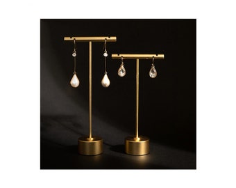 May include: A gold metal earring stand with two pairs of earrings. The stand has a round base and a T-shaped top. One pair of earrings is white and the other pair is clear.