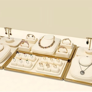 Pode incluir: Uma coleção de joias exibidas em suportes de cor creme com molduras de metal dourado. O conjunto inclui um colar de pedras preciosas multicoloridas, vários anéis dourados, pulseiras e pares de brincos com pedras escuras e claras.