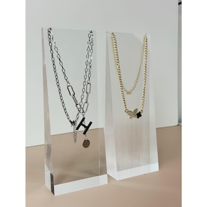 May include: Two necklaces displayed on clear acrylic stands. One is silver-toned with a layered chain design and a pendant with the letter "H". The other is gold-toned with a butterfly pendant and a small black flower.