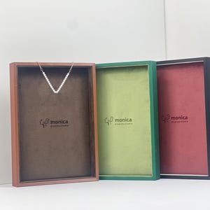 May include: Three rectangular display trays with wooden frames in brown, green, and black. Each tray features a velvet-like interior lining in brown, light green, or red, with the text "monica displaystudio" printed on the base. A white beaded necklace is draped over the brown tray.