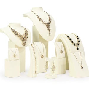 May include: A collection of cream-coloured jewellery display stands featuring various necklaces, bracelets, and earrings. The set includes multiple busts and pedestals showcasing gold-toned jewellery with clear, black, and metallic decorative accents.