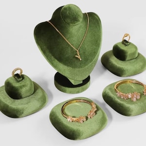 Könnte beinhalten: Eine Sammlung grüner Samt-Schmuckständer. Eine Halskette und mehrere Ringe und Armbänder werden auf den Ständern präsentiert. Der Schmuck ist goldfarben mit Blumen- und Buchstabenmotiven.