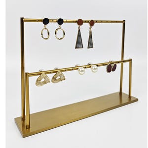 Könnte beinhalten: Ein goldfarbener Ohrring-Präsentationsständer aus Metall mit zwei horizontalen Stangen. Der Ständer hält verschiedene Ohrringe, darunter schwarze Ohrstecker, goldene Reifen sowie dreieckige und tropfenförmige Ohrringe in Braun- und Weißtönen.