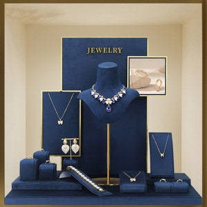 Puede incluir: Una exhibición de joyas, que incluye collares, aretes, pulseras y anillos, presentada en soportes y cajas de terciopelo azul. La palabra "JEWELRY" se muestra en letras doradas. Las joyas presentan diseños de mariposas y corazones, con detalles dorados y azules.