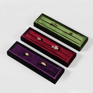 May include: Three rectangular velvet jewelry display boxes in green, red, and purple. Each box contains rings. The boxes are arranged diagonally on a white surface. The rings are gold and silver.
