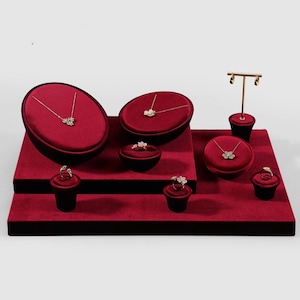 Op de afbeelding: Sieraden display met rode fluwelen platforms en zwarte sokkels. Verschillende sieraden, waaronder kettingen en ringen, worden getoond. De sieraden hebben een bloemenontwerp. Er is ook een goudkleurige oorbelstandaard aanwezig.