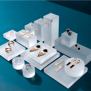 Puede incluir: Una colección de joyas que incluye anillos, collares y pendientes en tonos dorados y plateados expuestos sobre bloques de acrílico esmerilado. El conjunto presenta varios diseños como aros, pendientes colgantes con detalles negros.