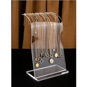 Peut inclure: Pr&eacute;sentoir transparent en acrylique pour colliers, pr&eacute;sentant plusieurs colliers dor&eacute;s. Les colliers ont diff&eacute;rents styles, avec des pendentifs en forme d'&eacute;toile, de pi&egrave;ce et de cercle. Le pr&eacute;sentoir a un dessus incurv&eacute; et une base rectangulaire.