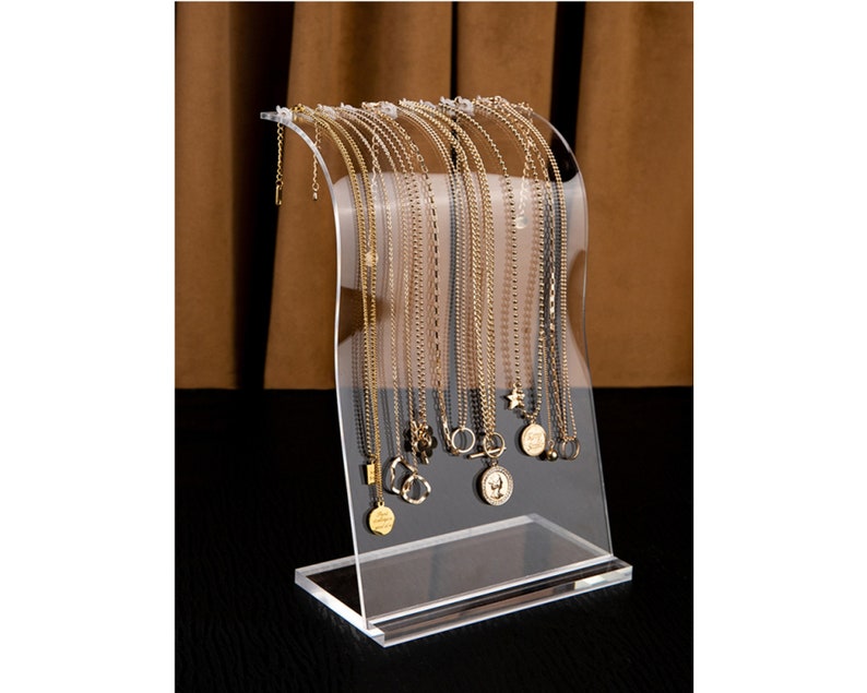 Peut inclure: Pr&eacute;sentoir transparent en acrylique pour colliers, pr&eacute;sentant plusieurs colliers dor&eacute;s. Les colliers ont diff&eacute;rents styles, avec des pendentifs en forme d'&eacute;toile, de pi&egrave;ce et de cercle. Le pr&eacute;sentoir a un dessus incurv&eacute; et une base rectangulaire.