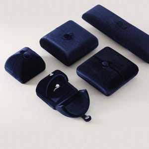 Può includere: Cinque scatole per gioielli in velluto blu. Una scatola è un rettangolo lungo, una è un quadrato e tre sono piccole e rotonde. Le scatole rotonde hanno coperchi che si aprono per rivelare un piccolo cuscino di velluto.