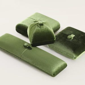 May include: A collection of four olive green velvet jewelry boxes in various shapes, including one long rectangular case, two square cases, and one dome-shaped case. Each box features a matching velvet button closure on the top center.