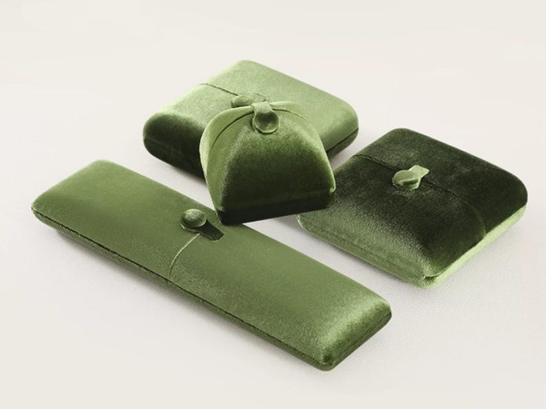 May include: A collection of four olive green velvet jewelry boxes in various shapes, including one long rectangular case, two square cases, and one dome-shaped case. Each box features a matching velvet button closure on the top center.