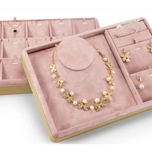Puede incluir: Una caja de presentación de joyas de terciopelo rosa con ribete dorado. La caja tiene un soporte para collar y varios compartimentos para pendientes, anillos y collares.