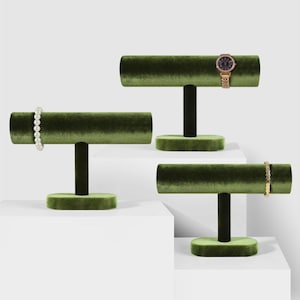 May include: Three olive green velvet jewelry display stands. Each stand has a cylindrical top, a black stem, and a rounded base. A pearl bracelet, a gold watch, and a gold bracelet are displayed on the stands.