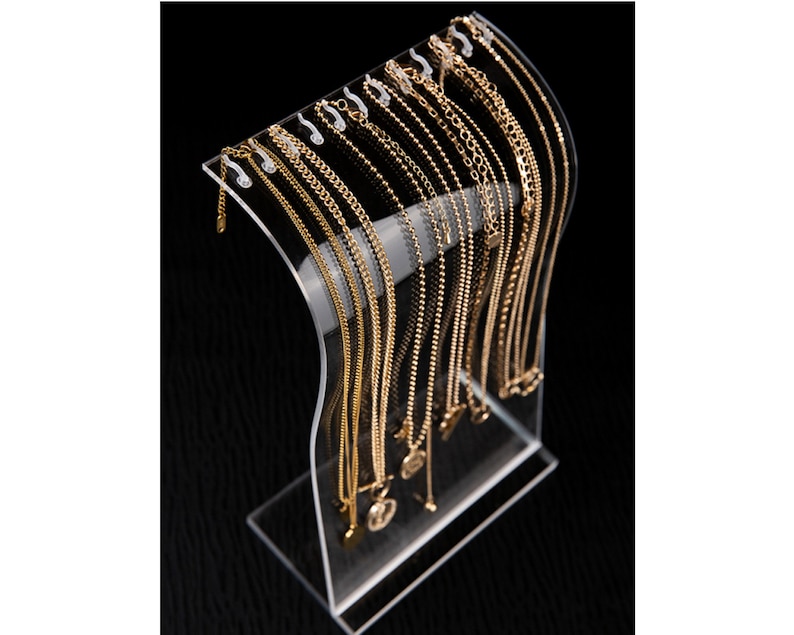 Peut inclure: Un pr&eacute;sentoir &agrave; bijoux en acrylique transparent pr&eacute;sentant plusieurs colliers dor&eacute;s. Les colliers varient en style, y compris des mod&egrave;les &agrave; cha&icirc;ne et &agrave; perles. Le pr&eacute;sentoir a un design incurv&eacute; et une base robuste.