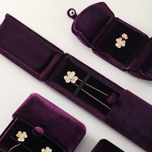 Op de afbeelding: Een bovenaanzicht van sieraden gepresenteerd in open, dieppaarse fluwelen etuis. De sieraden omvatten een gouden ketting en oorbellen, elk met een klavervormig ontwerp en sprankelende details. De etuis zijn gerangschikt op een licht oppervlak.