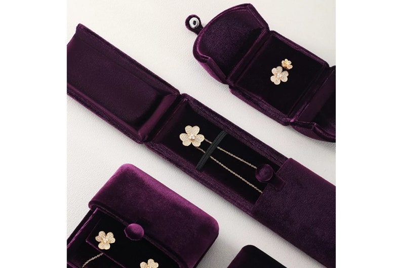 Op de afbeelding: Een bovenaanzicht van sieraden gepresenteerd in open, dieppaarse fluwelen etuis. De sieraden omvatten een gouden ketting en oorbellen, elk met een klavervormig ontwerp en sprankelende details. De etuis zijn gerangschikt op een licht oppervlak.