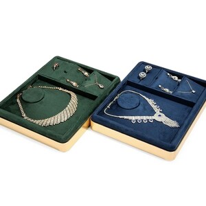 May include: Two rectangular jewelry display trays with gold-toned metal frames. One tray features a dark green velvet interior, the other a navy blue velvet interior. Both contain compartments holding necklaces, earrings, and small jewelry pieces.