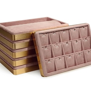 Könnte beinhalten: Ein Stapel rosa Samt-Schmuck-Display-Tabletts mit goldfarbenem Metallbesatz. Ein Tablett ist geöffnet und zeigt einzelne Fächer für Ohrringe oder kleine Gegenstände. Die Tabletts dienen zum Ordnen und Präsentieren von Schmuck.