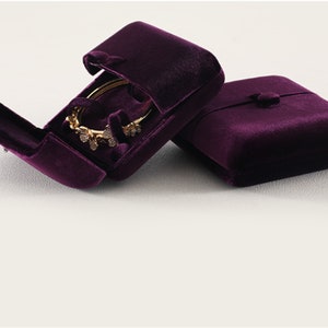 Op de afbeelding: Twee pruimkleurige fluwelen sieradendoosjes, &eacute;&eacute;n open en &eacute;&eacute;n gesloten. Het open doosje toont een gouden armband met decoratieve versieringen. De doosjes hebben een knoopsluiting en zijn ontworpen om sieraden te beschermen en te presenteren.