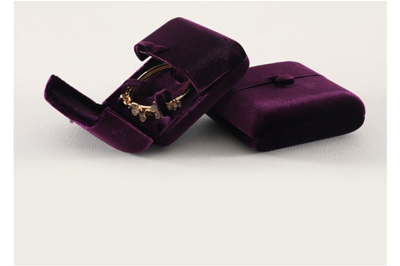 Op de afbeelding: Twee pruimkleurige fluwelen sieradendoosjes, &eacute;&eacute;n open en &eacute;&eacute;n gesloten. Het open doosje toont een gouden armband met decoratieve versieringen. De doosjes hebben een knoopsluiting en zijn ontworpen om sieraden te beschermen en te presenteren.