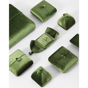 May include: A collection of olive green velvet jewelry boxes in various square and rectangular shapes, featuring button closures. One open box in the center displays a small gold ring inside, while the other boxes remain closed.