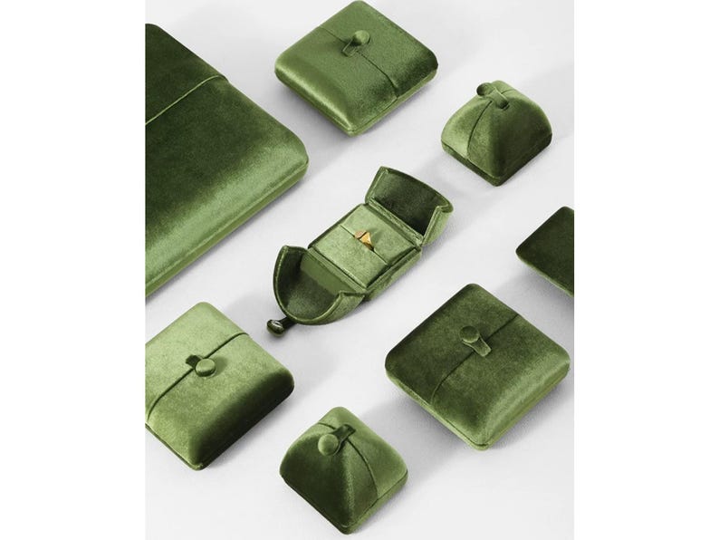 May include: A collection of olive green velvet jewelry boxes in various square and rectangular shapes, featuring button closures. One open box in the center displays a small gold ring inside, while the other boxes remain closed.