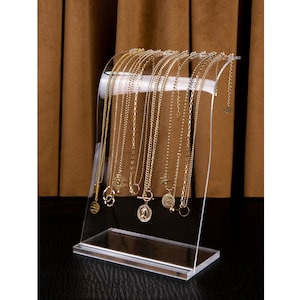 Peut inclure: Pr&eacute;sentoir &agrave; colliers en acrylique transparent pr&eacute;sentant plusieurs colliers dor&eacute;s avec diff&eacute;rents mod&egrave;les de pendentifs. Le pr&eacute;sentoir a un design incurv&eacute; et une base rectangulaire, id&eacute;al pour les bijoux.